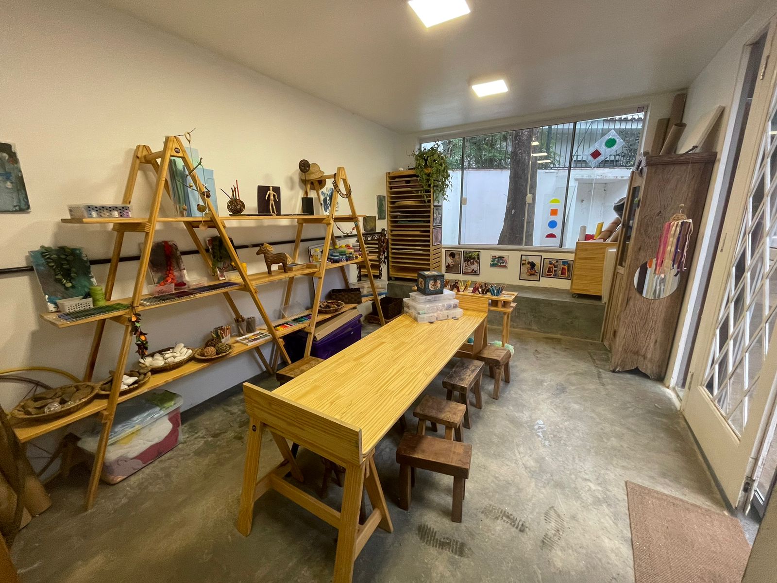Ateliê com mesa de madeira, banquinhos e prateleiras de materiais