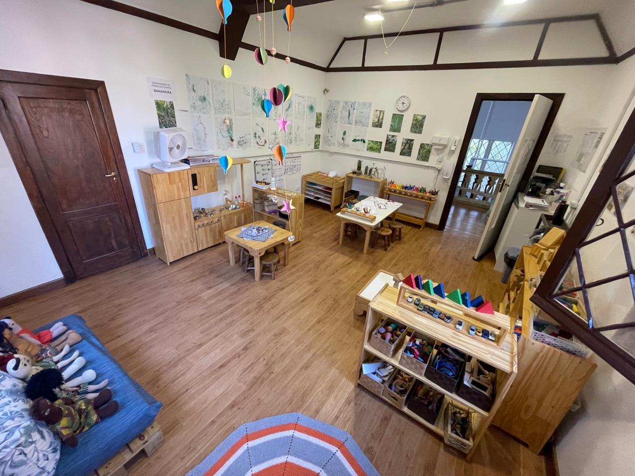 Sala de referência com cozinha de madeira, mesas baixas e painel de desenhos das crianças